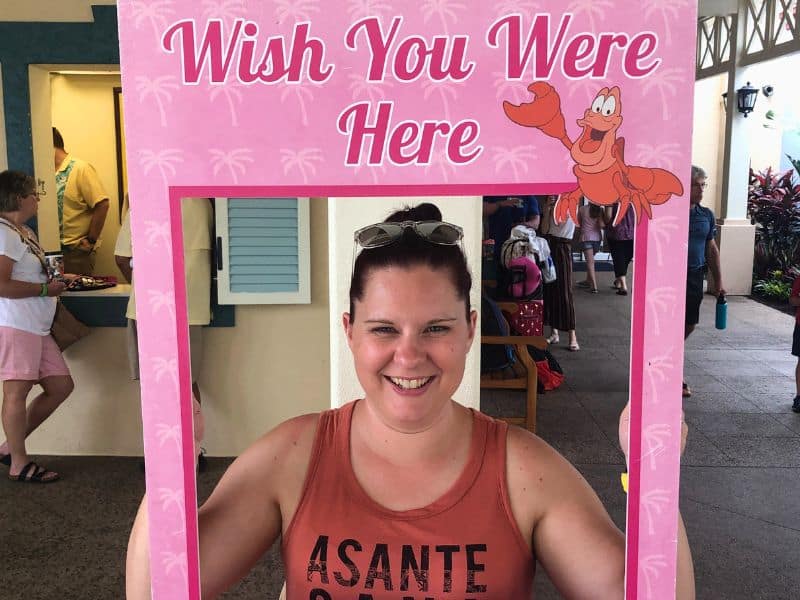 Woman smiling while holding up a sign that says "Wish you were here - Disney's Caribbean Beach Resort."