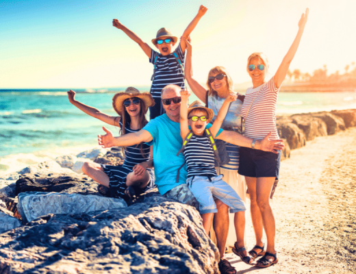A photo of a Multi-generation family vacation, with everyone holding their hands up and smiling by beach.