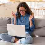 A woman sitting on the couch, in front of her computer, looking very excited.